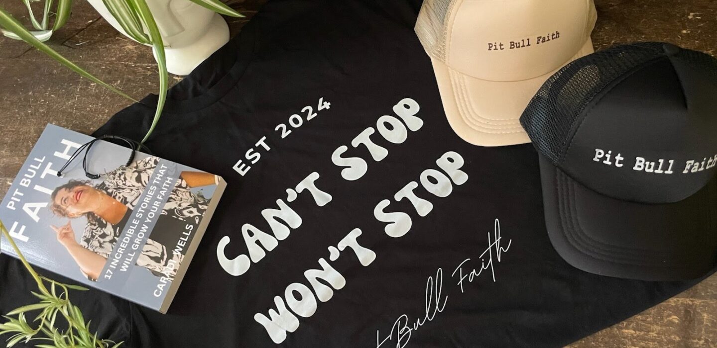 A Pit Bull Faith branded "Can't Stop Won't Stop" black t-shirt and hat displayed on a wooden surface.